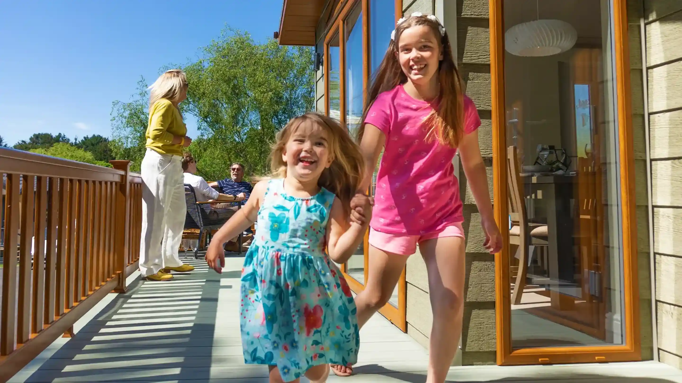 Two girls joyfully run along a wooden deck, holding hands. One girl wears a blue floral dress and the other a pink shirt and shorts. In the background, a woman in a yellow top and a group of people can be seen relaxing. The sky is clear and sunny.