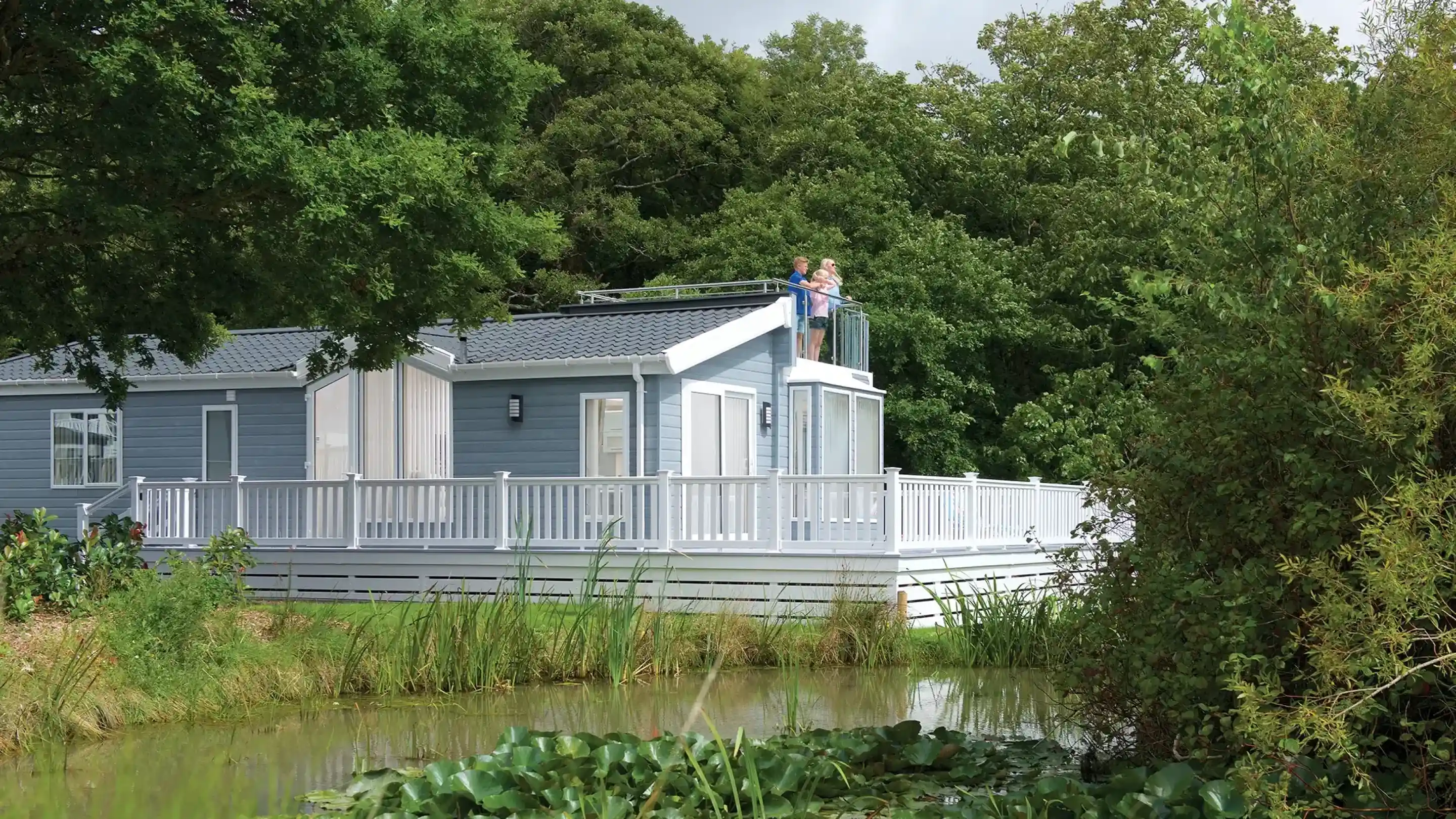 A modern blue house with a spacious deck overlooks a serene pond surrounded by lush greenery. Two people stand on the deck, enjoying the view.