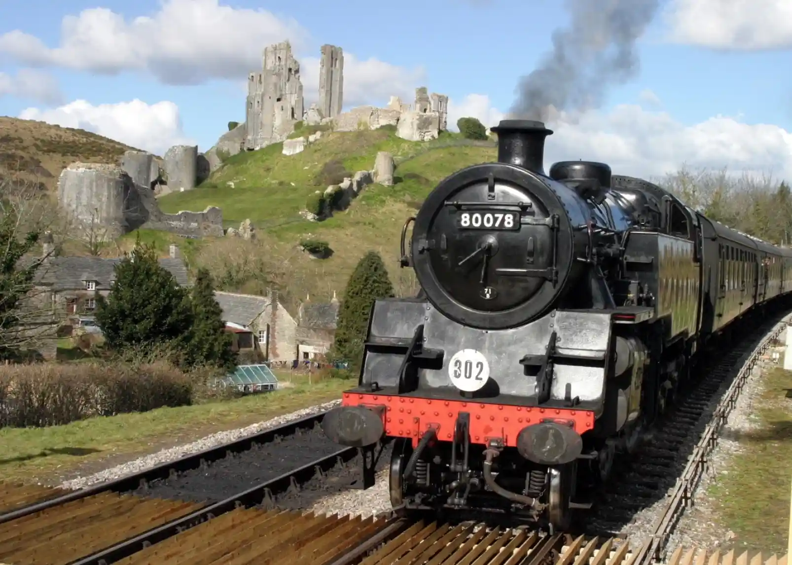 A vintage steam locomotive travels along tracks in the foreground, with a backdrop of a historic castle and rolling hills under a partly cloudy sky.