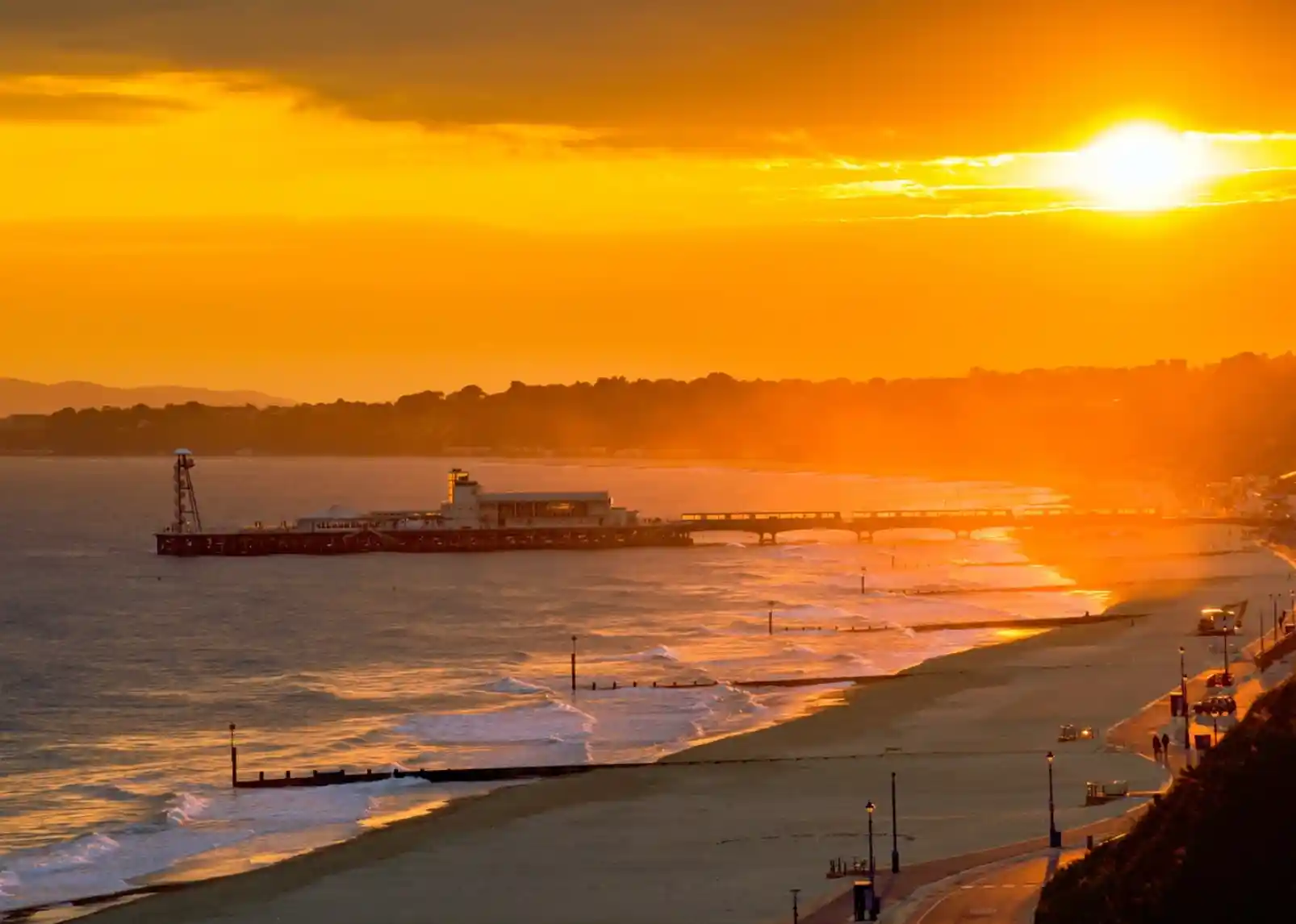 A vibrant sunset casts golden hues over a beach, with a pier extending into the water. The coastline is lined with lights and gentle waves lap at the shore.