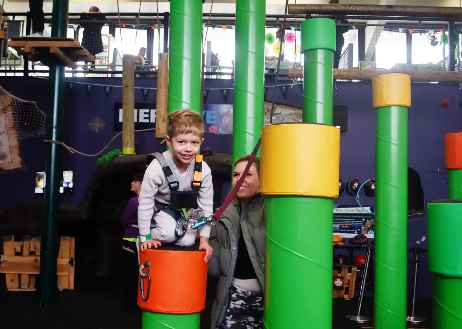 A young boy climbing on colourful cylindrical structures in an indoor playground, with a woman watching and providing support nearby.
