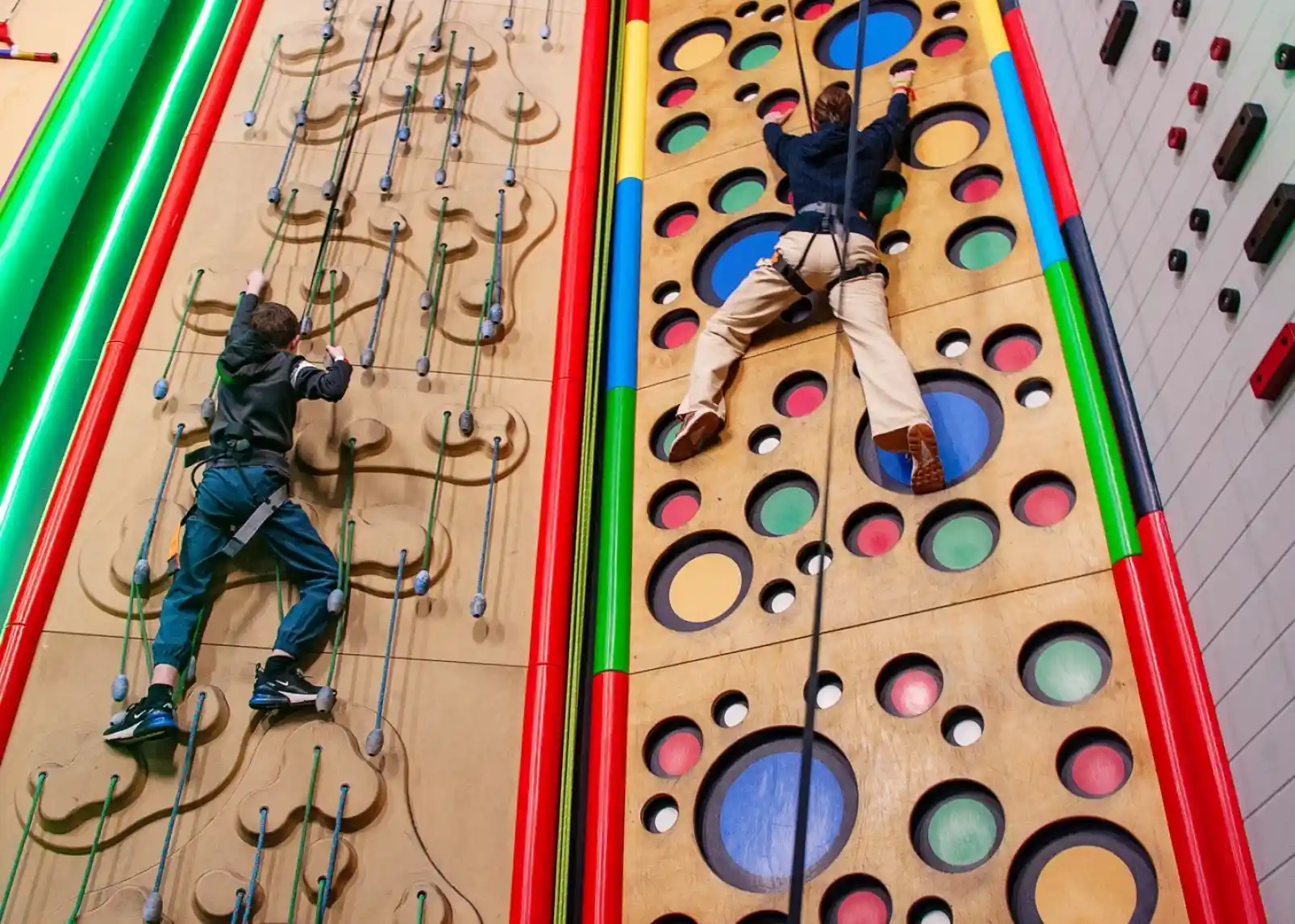 Two climbers scaling a colourful indoor climbing wall. One climber ascends a wall with grips shaped like circles, while the other uses a more textured surface with climbing holds. Bright colours and bold patterns enhance the vibrant atmosphere of the climbing gym.