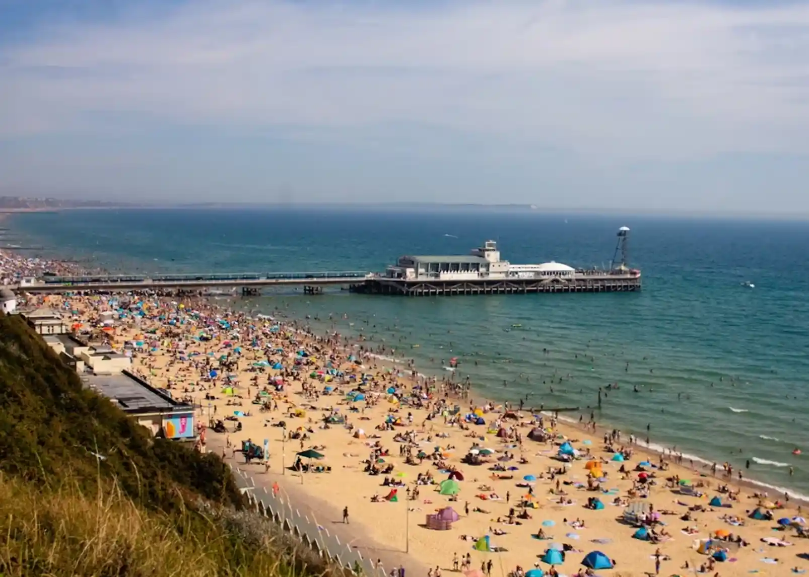 A busy beach scene with colourful umbrellas and sunbathers on the sand, stretching towards a wooden pier extending into the turquoise sea. Clear blue skies are overhead, and people are enjoying the water and the beach activities.
