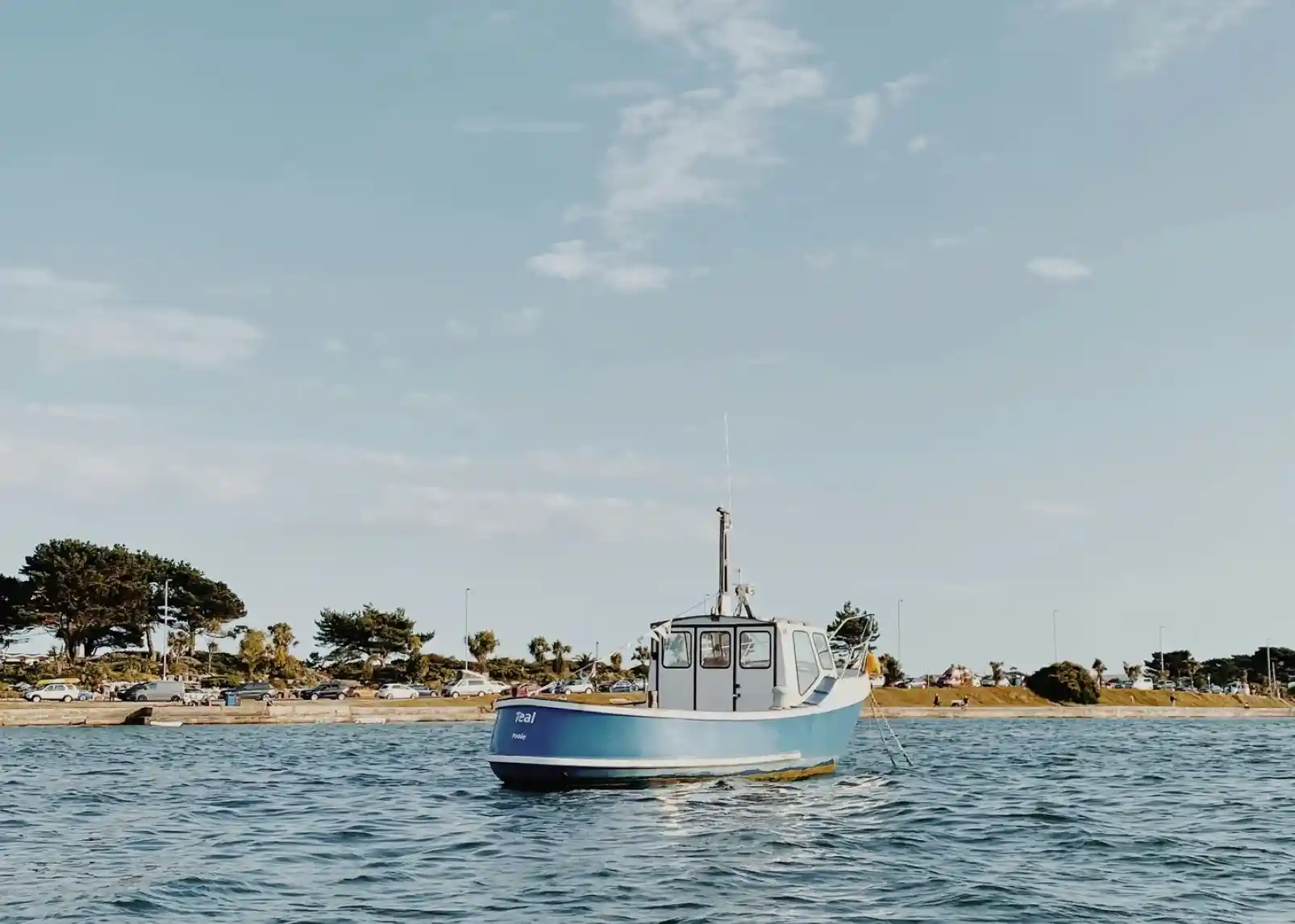 A small blue fishing boat floats on calm waters, with a scenic shoreline and trees in the background under a clear sky.