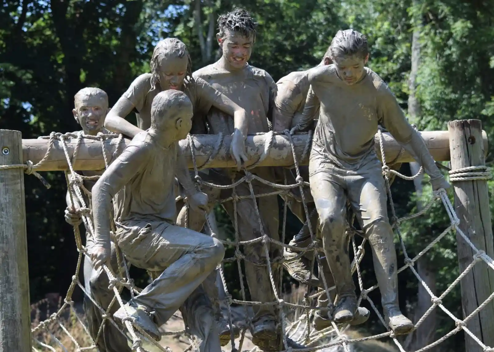 Children covered in mud play on a rope climbing structure, laughing and interacting as they climb and hang. Sunlight filters through trees in the background, creating a lively outdoor scene.