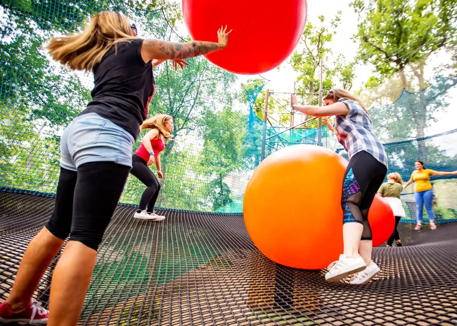 People playing on a large, netted trampoline surrounded by colourful, oversized balls in a vibrant outdoor setting. Trees and blue sky are visible in the background, creating a lively atmosphere.