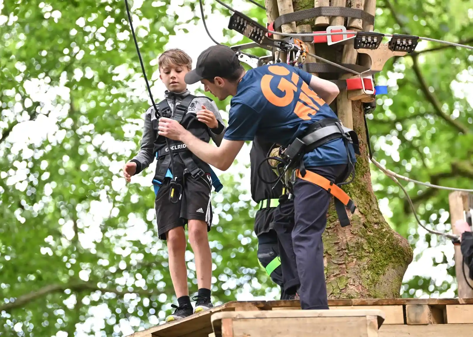 A young boy in a black outfit receives guidance from an instructor while standing on a wooden platform in a tree-top adventure park. The background features lush green trees, creating a vibrant outdoor setting.