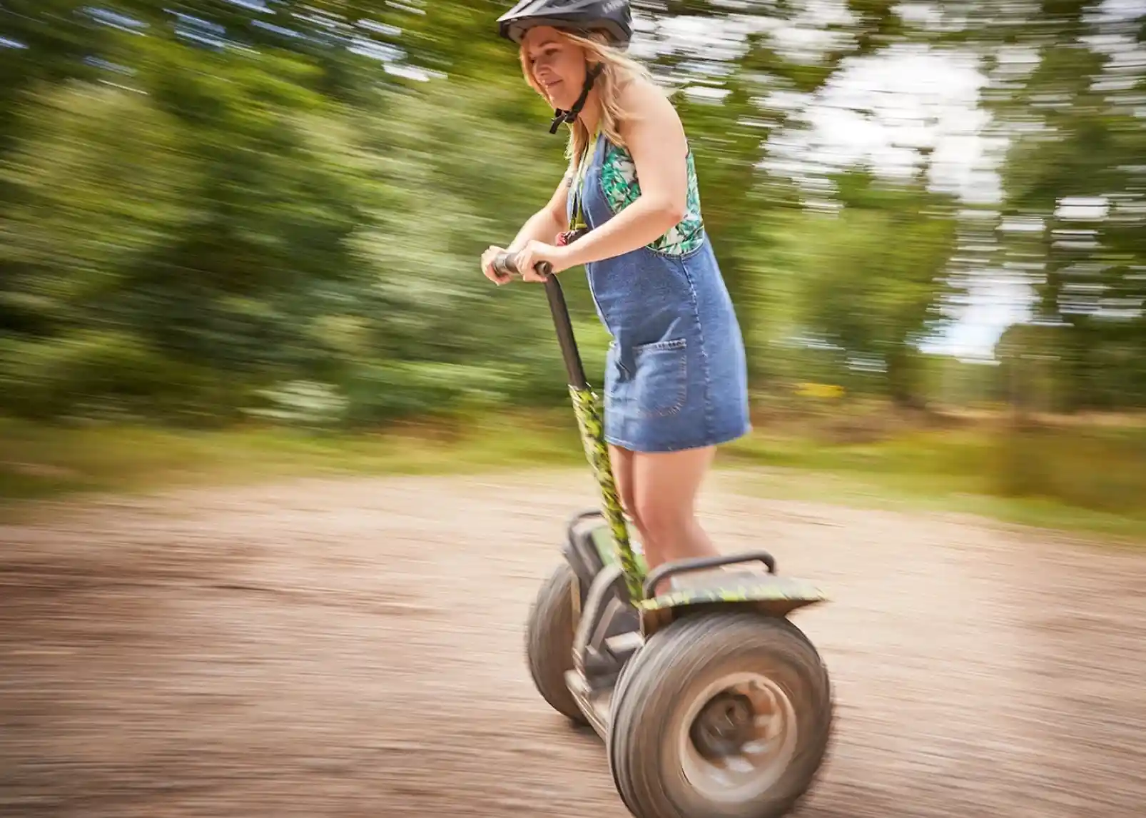 A young woman rides a scooter on a gravel path, wearing a helmet and a colourful top. The background is blurred, conveying a sense of motion.