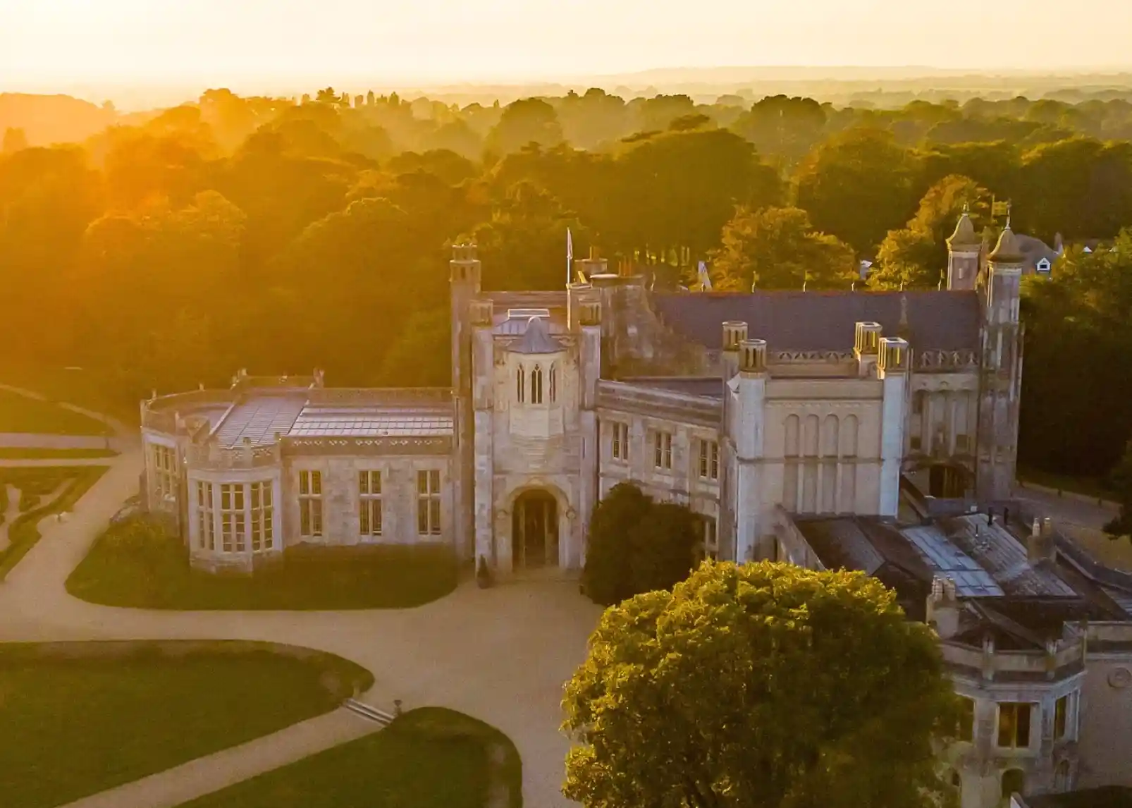 A grand historic mansion surrounded by lush trees, illuminated by a warm golden sunset. The architecture features turrets and detailed stonework, with pathways winding around the property. Photo credit -Andrew Millard