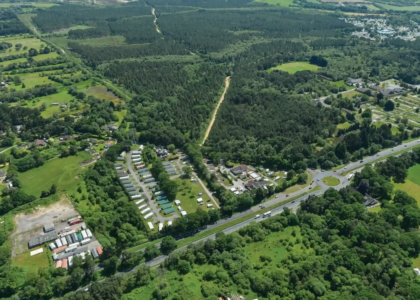 Bird's-eye view of a green landscape featuring a mix of forests, fields, and a residential area with a caravan park. A road runs along the bottom, connecting to the nearby village and surrounding rural scenery.