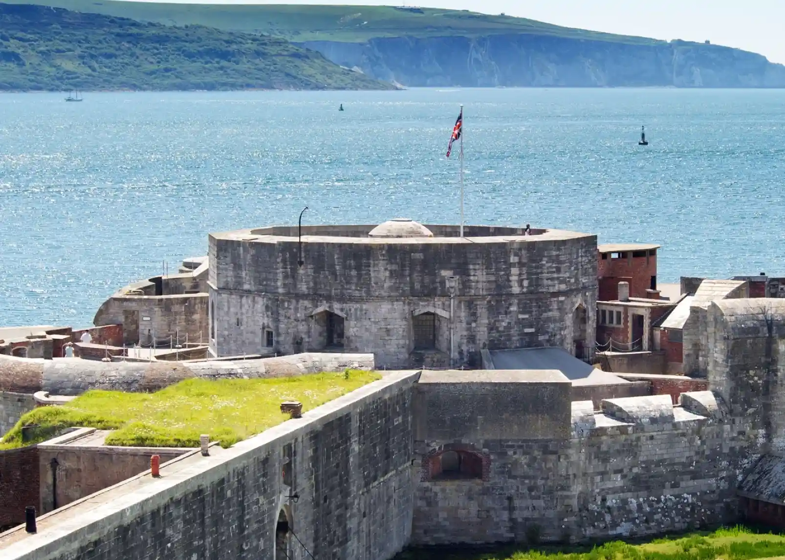 A historic stone fort with a circular tower and a flag flying at the top overlooks a calm blue sea. In the background, green hills rise against a clear sky, dotted with small boats.