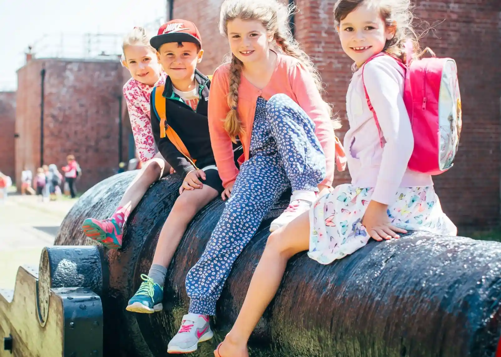 Four children, two girls and two boys, sit on a large cannon in a sunny outdoor setting. They wear colorful clothing, and backpacks are visible on two of the children. A brick wall and other kids can be seen in the background.