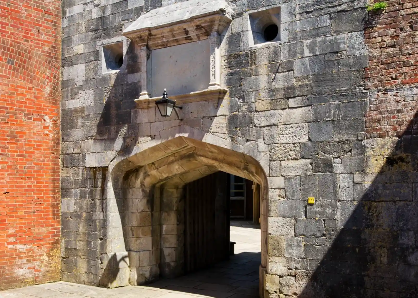 A historic stone archway surrounded by brick walls, featuring a decorative ledge above the entrance and a vintage lantern hanging beside it. The ground is paved with stone slabs, leading into a courtyard area.