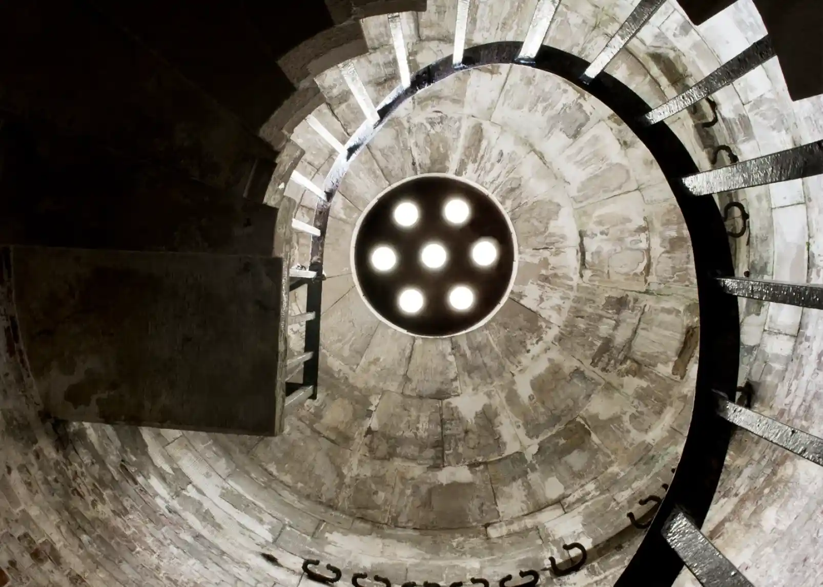 A spiral staircase leads upward, featuring a circular light aperture at the top. The walls are made of aged stone, and metal handrails outline the steps.
