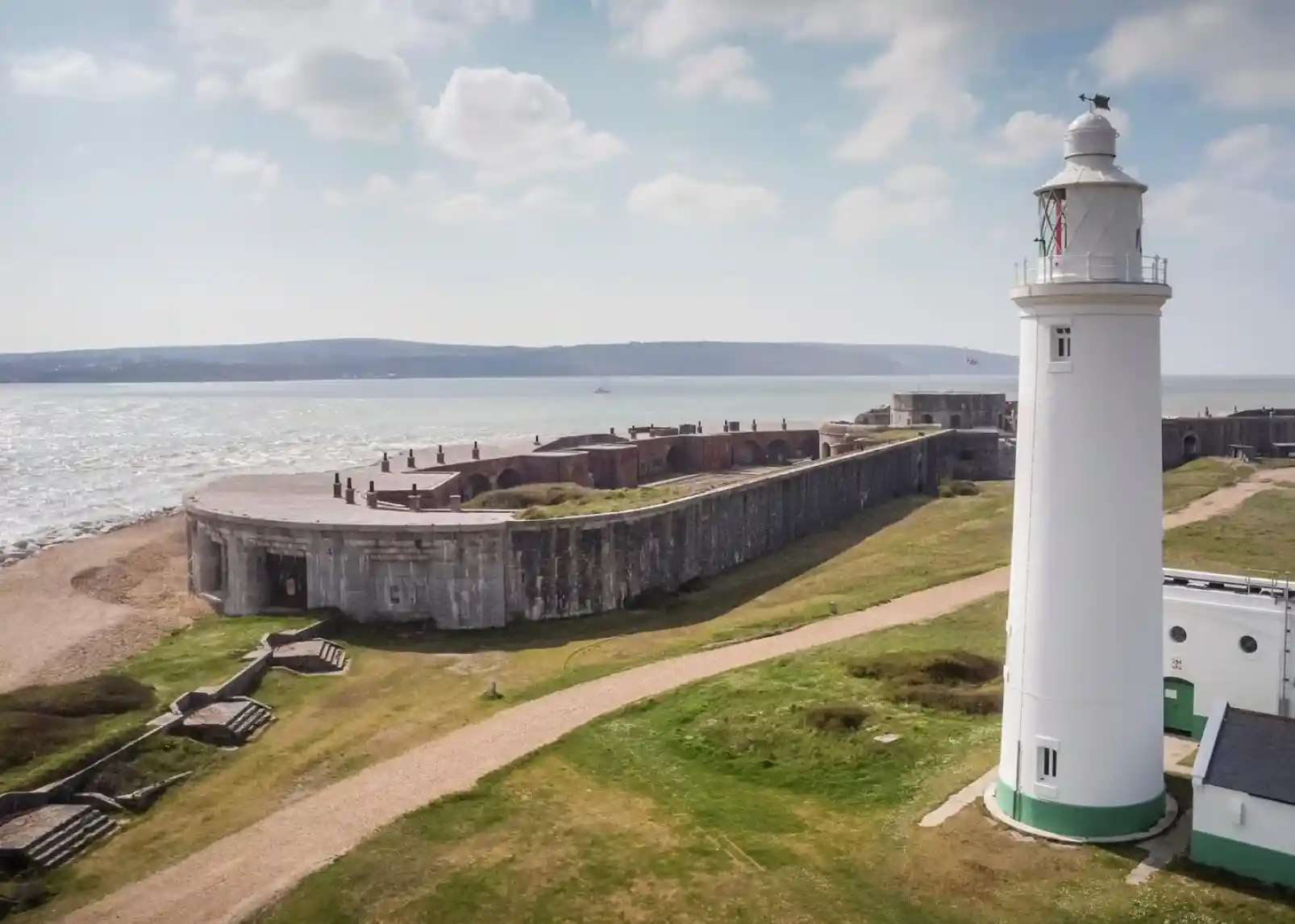 A white lighthouse stands near a historic fort overlooking a calm sea. Lush green grass and sandy pathways surround the structures, with clouds scattered across the blue sky.
