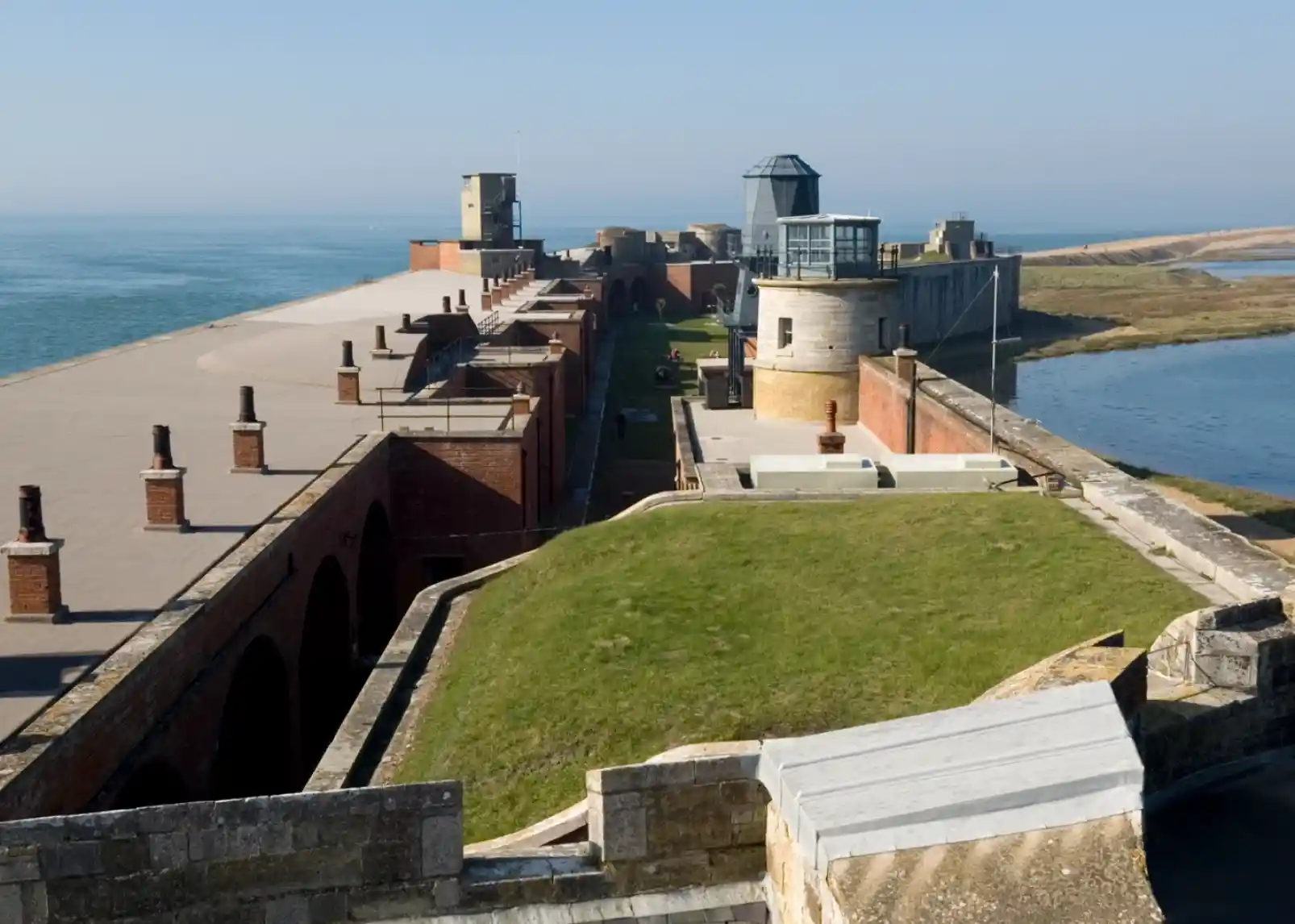 A historic coastal fortification with stone and brick structures along the shoreline, featuring grassy areas, walkways, and a blue sea in the background.