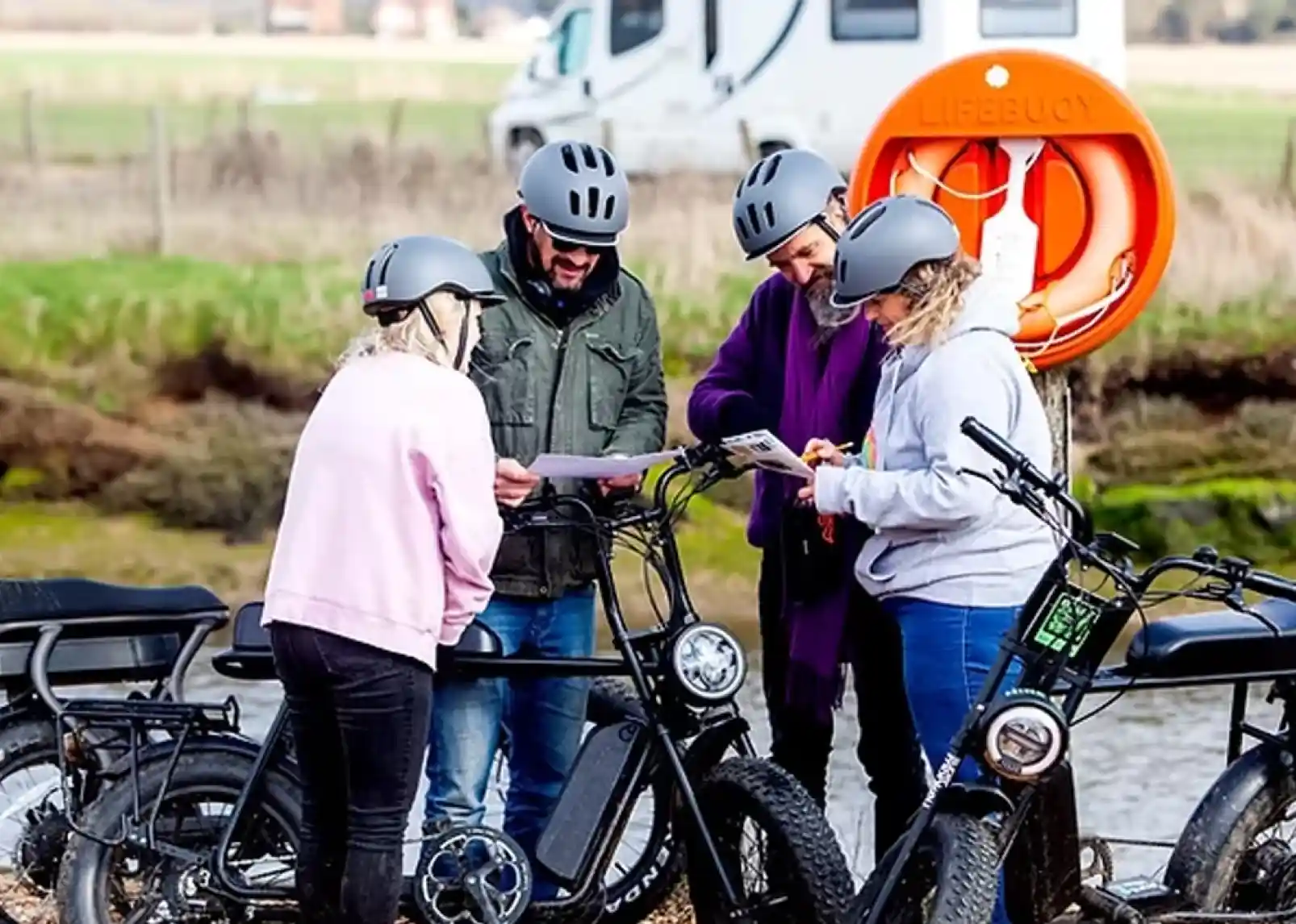 A group of four people wearing helmets gathers around electric bikes while looking at a map. They are standing near a body of water, with a lifebuoy visible in the background and a recreational vehicle parked nearby.