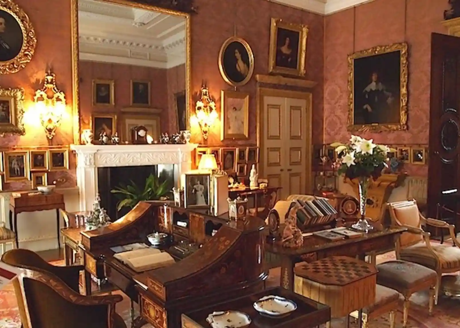 A lavishly decorated room featuring intricate wallpaper, ornate gold-framed portraits, a fireplace adorned with decorative items, and a mix of elegant furniture including a wooden writing desk. Various displayed artworks and floral arrangements add to the opulent atmosphere.