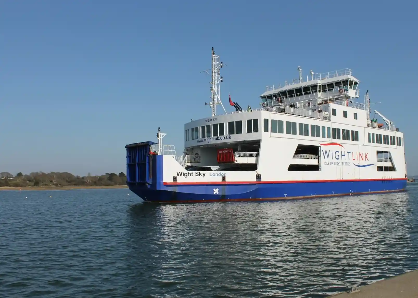 A large ferry named "Wight Sky" is docked at a port under a clear blue sky. The ferry features a white and blue color scheme with the Wightlink logo prominently displayed. Surrounding the vessel are calm waters and a distant shoreline.