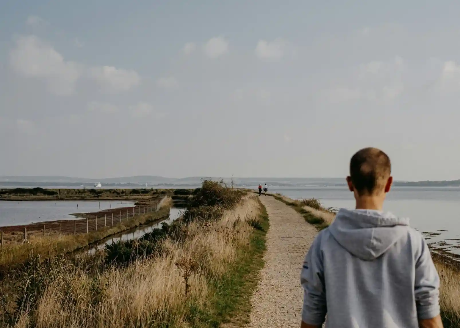A person walking along a gravel path by a body of water, with fields on either side and distant hills under a cloudy sky.