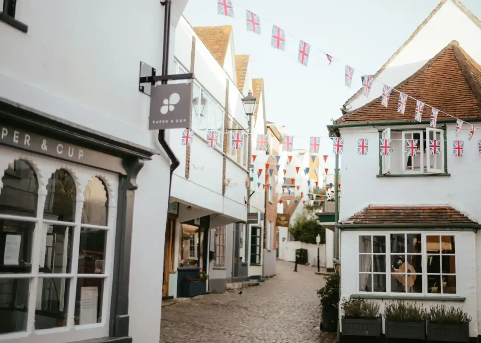 A quaint, cobblestone street lined with charming white buildings. Colorful bunting hangs overhead, featuring Union Jack and triangular flags. A sign reading "Paper & Cup" is visible on the left.