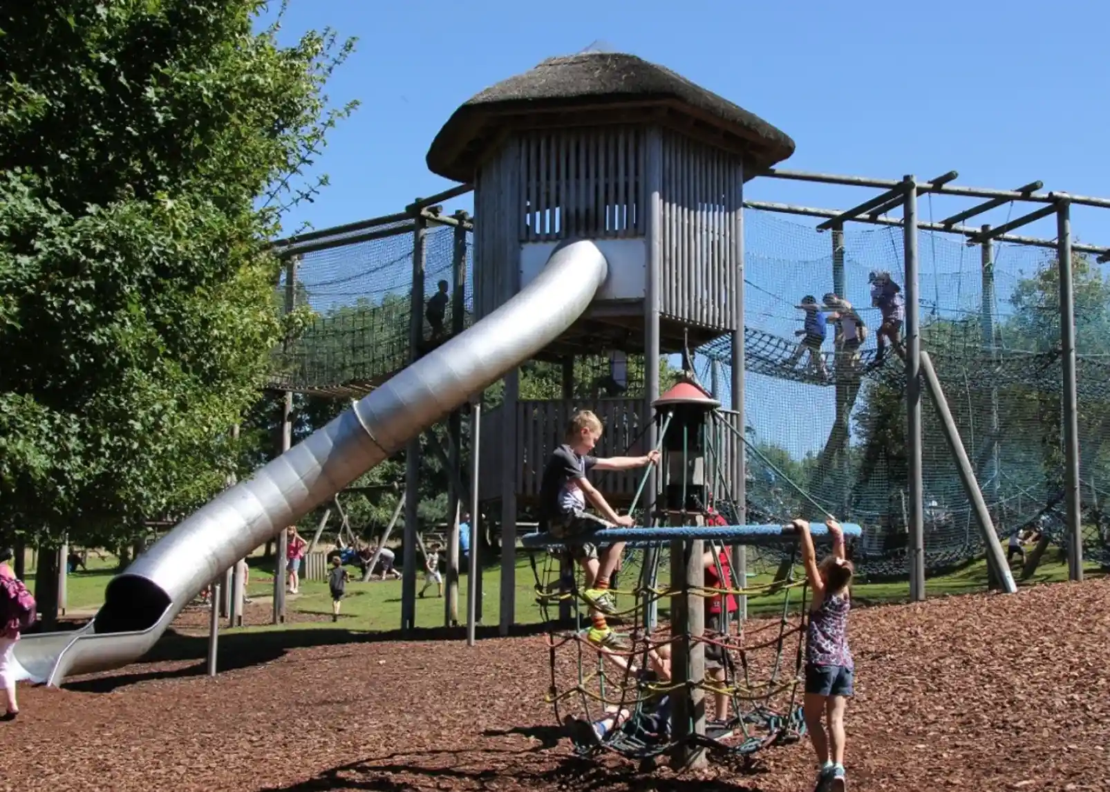 A wooden playground structure with a tall slide and netted climbing areas. Two children play on a spinning climbing apparatus in the foreground, while others are climbing on the structure above under clear blue skies. Lush green trees surround the area.