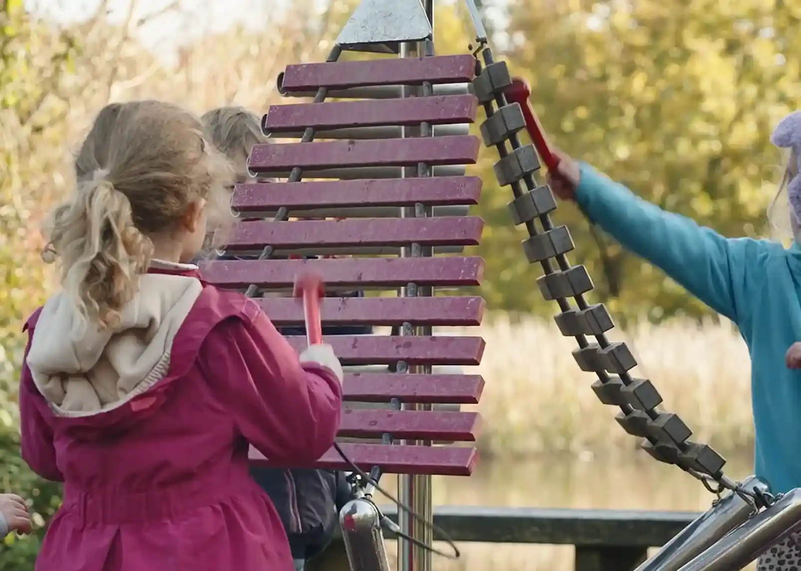 Children playing on a musical playground instrument with wooden slats and metal tubes, surrounded by trees and a body of water in the background.