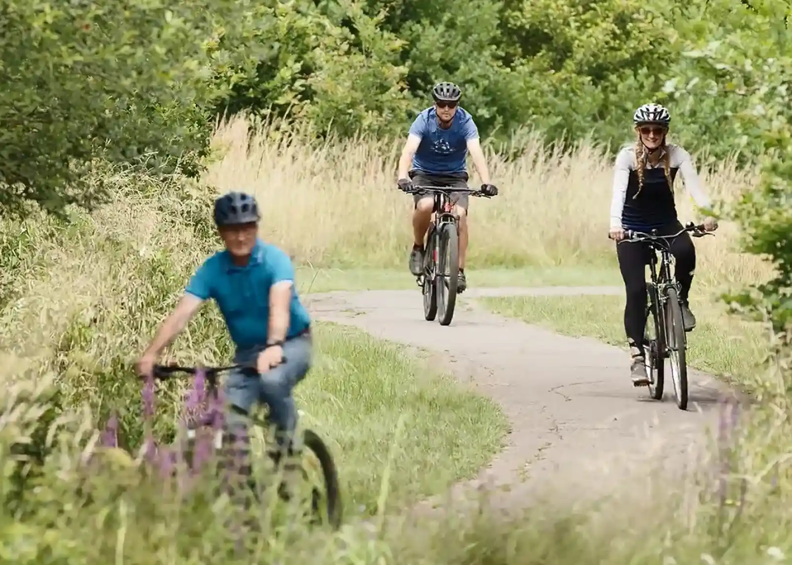 Three cyclists ride along a winding path in a lush green landscape. Two wear helmets and casual athletic clothing, while one cyclist, in a blue shirt, rides ahead on a bike. Tall grass and wildflowers line the path, creating a vibrant outdoor scene.