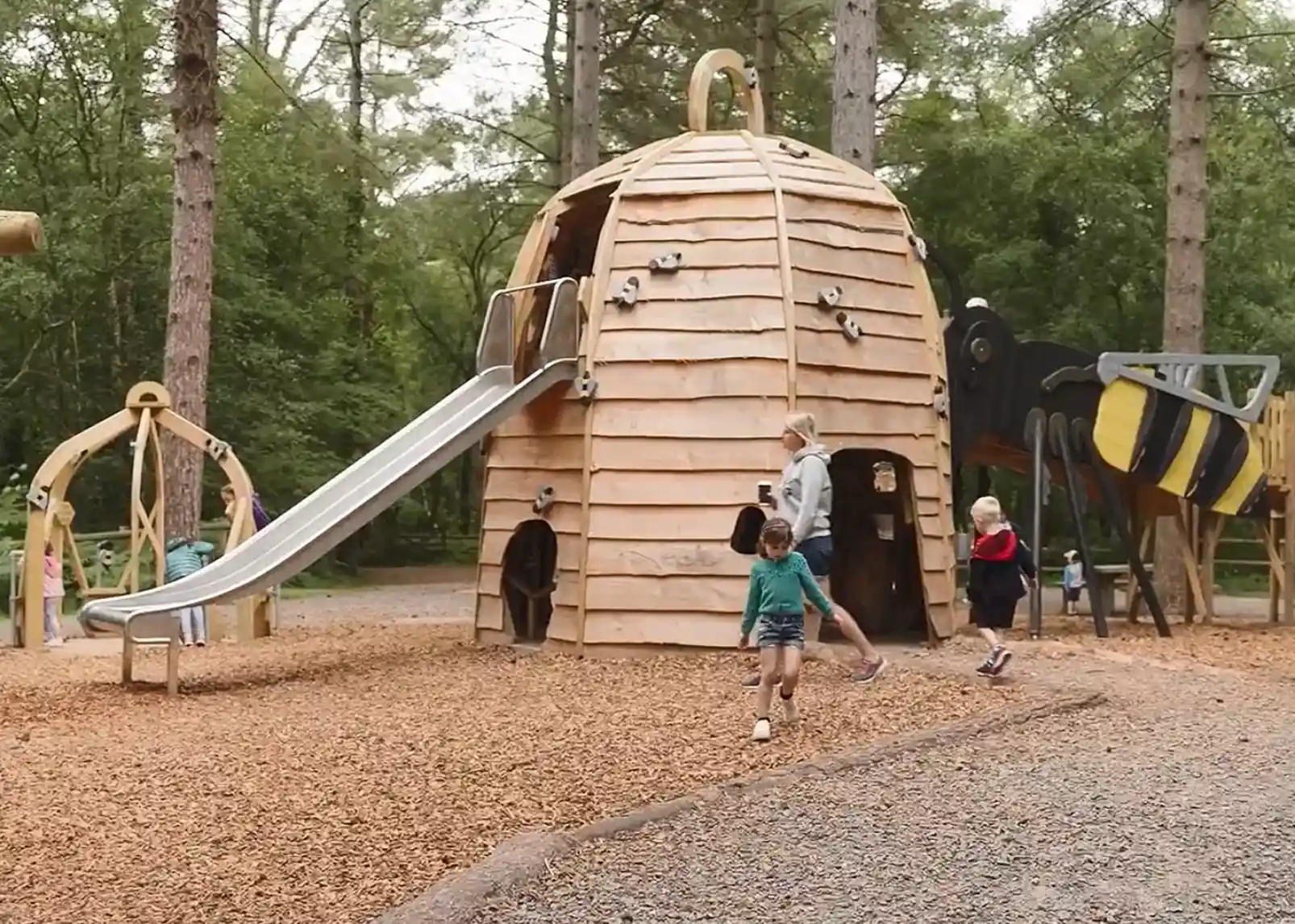 A wooden playground structure resembling a beehive, featuring a slide and climbing elements. Children play around the area, with some exploring the playground and others running on the gravel surface. Tall trees surround the play area, creating a natural setting.