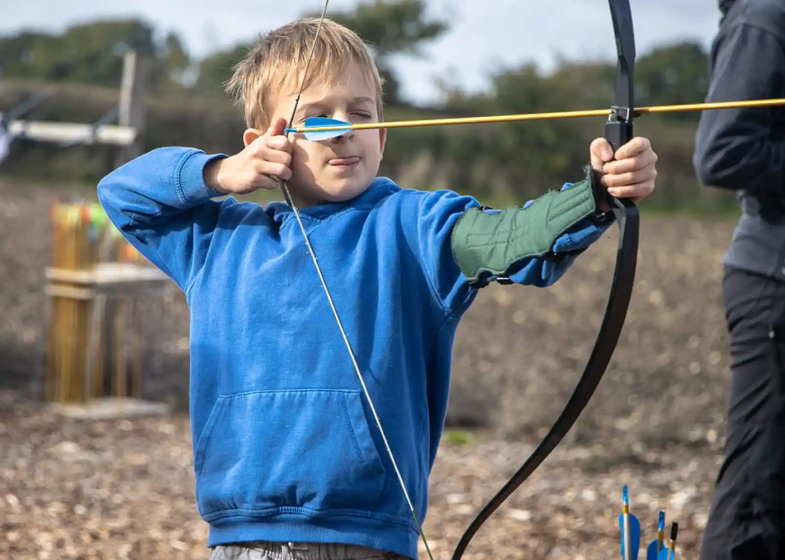 A young boy in a blue hoodie draws a bow and aims at a target with a focused expression. In the background, arrows can be seen in a target area, and another person is standing nearby.
