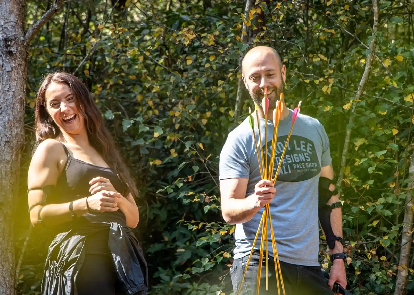 A man and a woman laugh while standing in a wooded area. The man holds several brightly colored arrows, and the woman has her arms crossed, smiling at him. Sunlight filters through the trees, illuminating their joyful expressions.