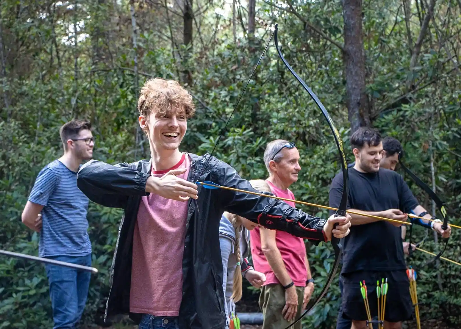 A young man with curly hair smiles while drawing a bow, surrounded by others in a forest setting. They are participating in an archery activity, with several arrows seen in the background.