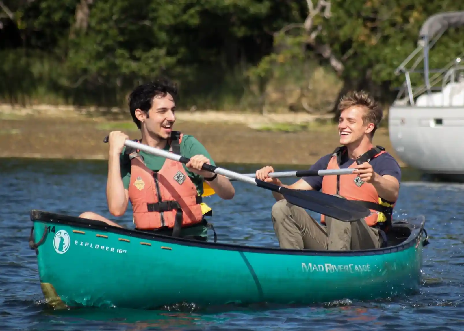 Two young men paddling together in a green canoe on a calm body of water. Both are wearing orange life jackets and smiling at each other, enjoying a sunny day outdoors. In the background, trees and a boat are visible.