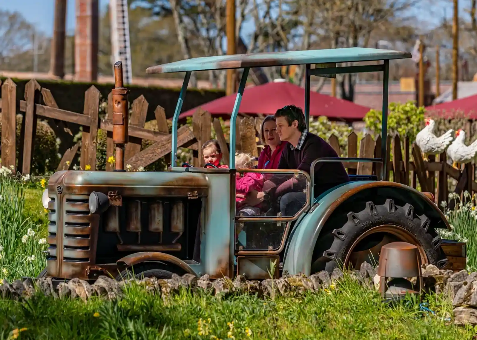 A family sits on a vintage tractor in a garden filled with flowers. Two adults and a child are smiling, with the child wearing sunglasses. In the background, there are wooden fences and red umbrellas.