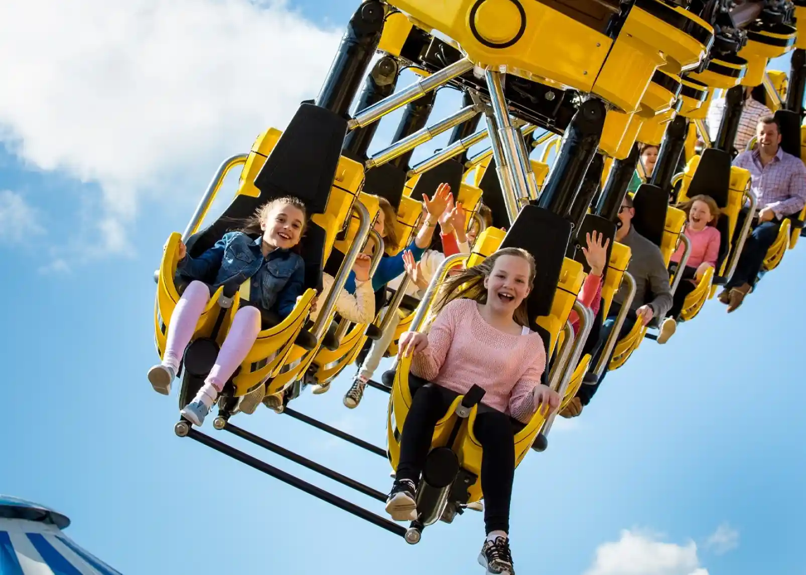 A group of children and adults ride a thrilling amusement park attraction, smiling and raising their hands in excitement against a bright blue sky with scattered clouds.