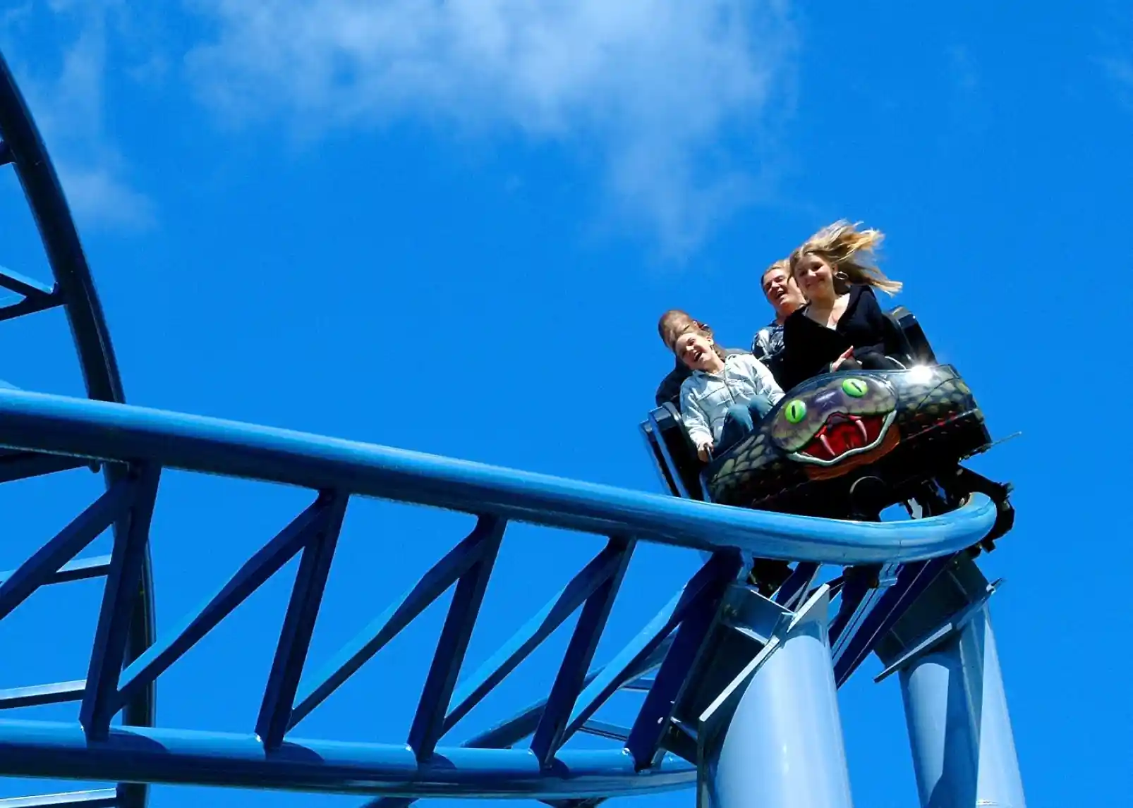 Three people are enjoying a thrilling ride on a roller coaster, with their hair blowing in the wind. The bright blue sky serves as a backdrop, highlighting the excitement of the moment.
