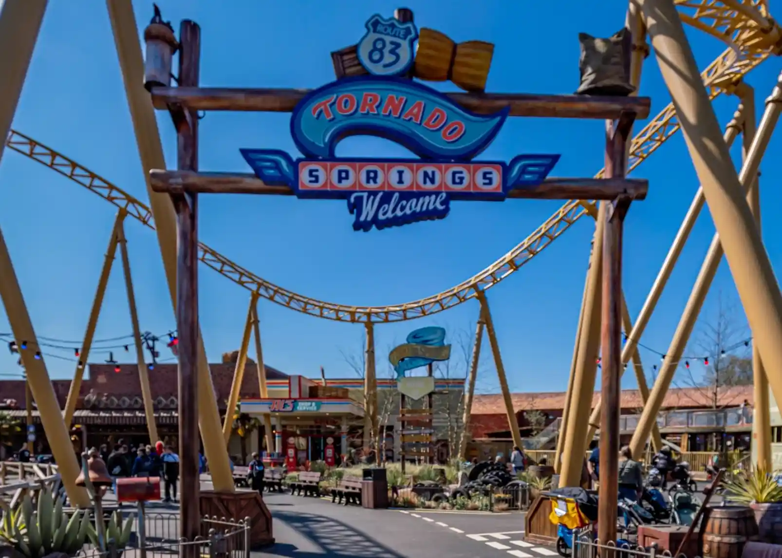 A brightly colored entrance sign welcomes visitors to Tornado Springs, featuring roller coaster tracks above and a lively theme park atmosphere with shops and attractions visible in the background.