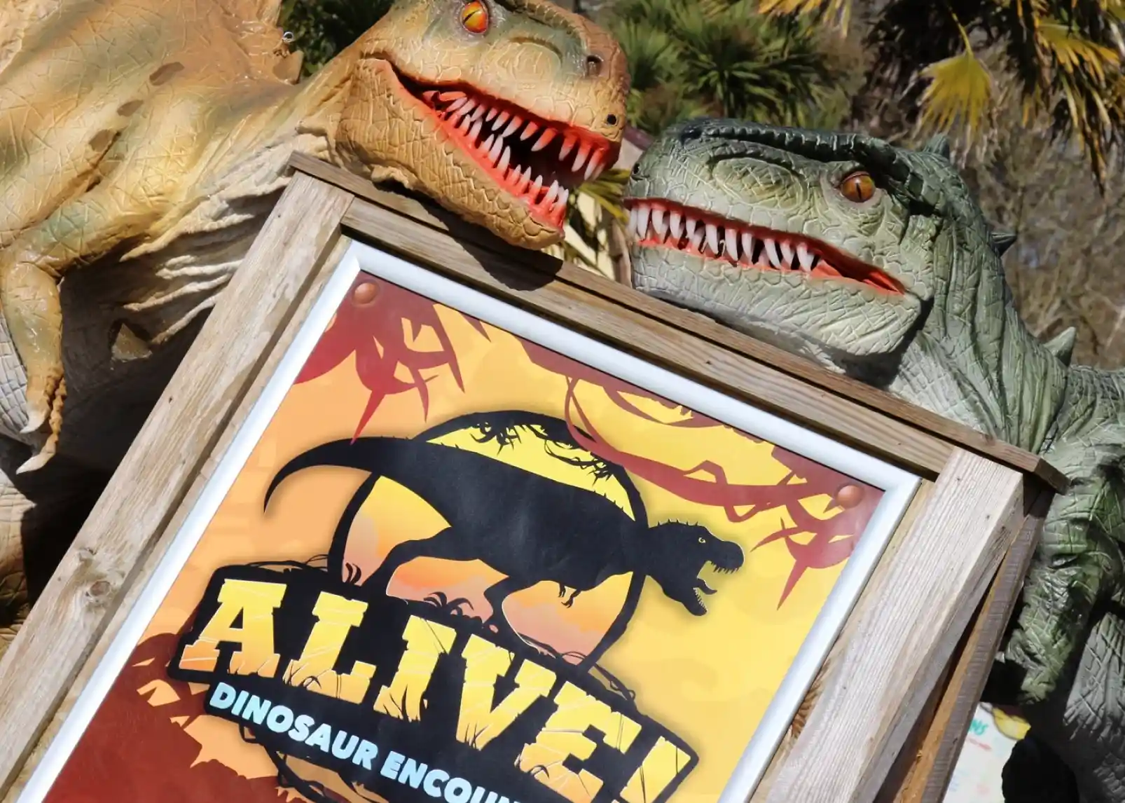 Two dinosaur sculptures with expressive faces stand on either side of a colorful sign that reads "Alive! Dinosaur Encounter," featuring a silhouetted dinosaur. Palm trees are visible in the background.