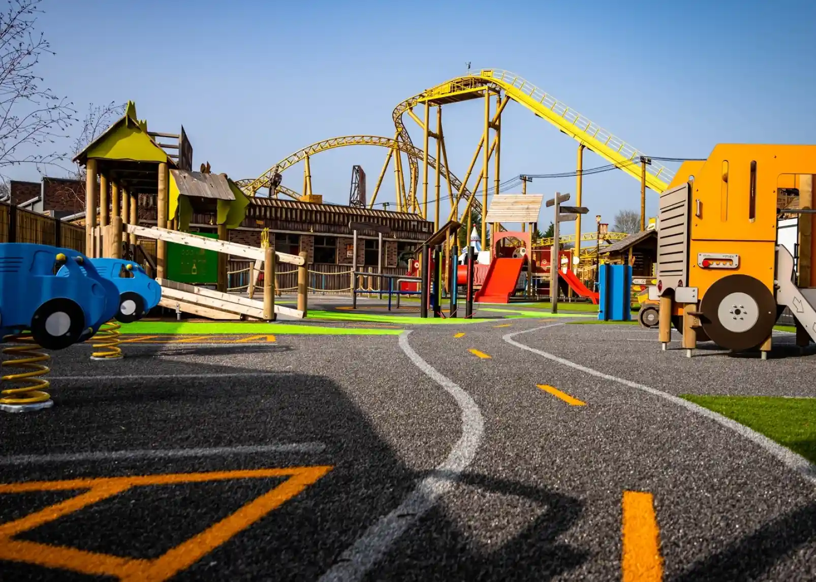 A colorful playground with a winding path features toy vehicles and a wooden play structure. In the background, a yellow roller coaster rises against a clear blue sky.