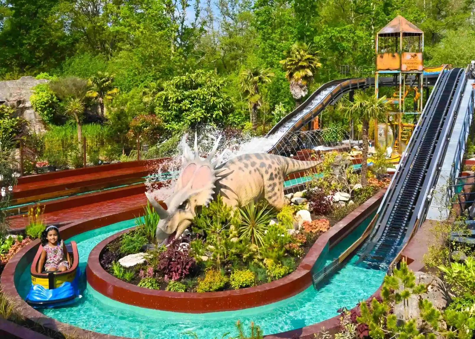 A colorful water slide attraction features a dinosaur sculpture in a lush, tropical setting. A child enjoys a ride in a small boat on the waterway, surrounded by vibrant plants and palm trees.