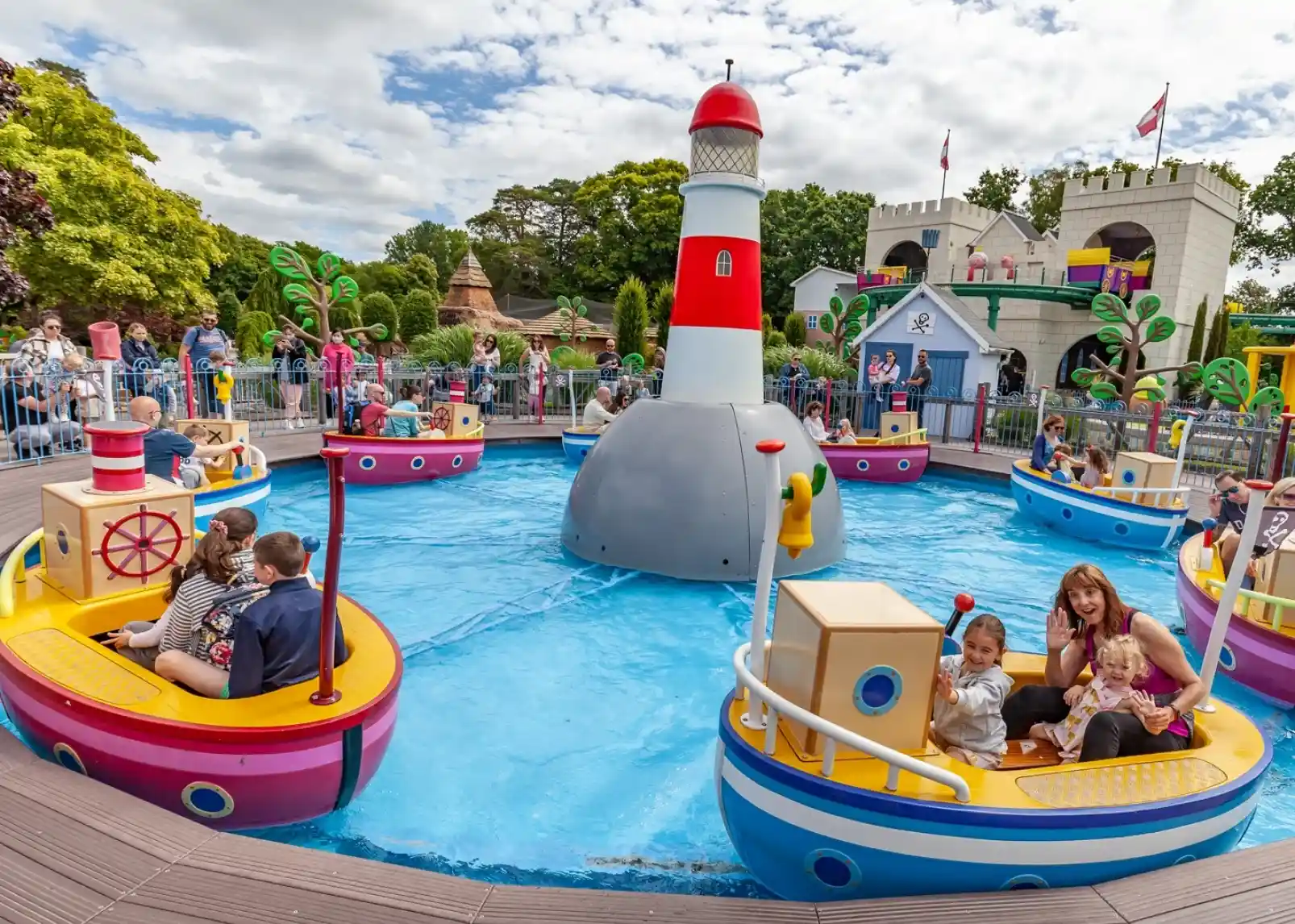 Colorful boats float in a playful, blue water area, surrounded by a cheerful lighthouse. Families enjoy the attractions in a vibrant amusement park, with trees and whimsical structures in the background. The sky is partly cloudy.