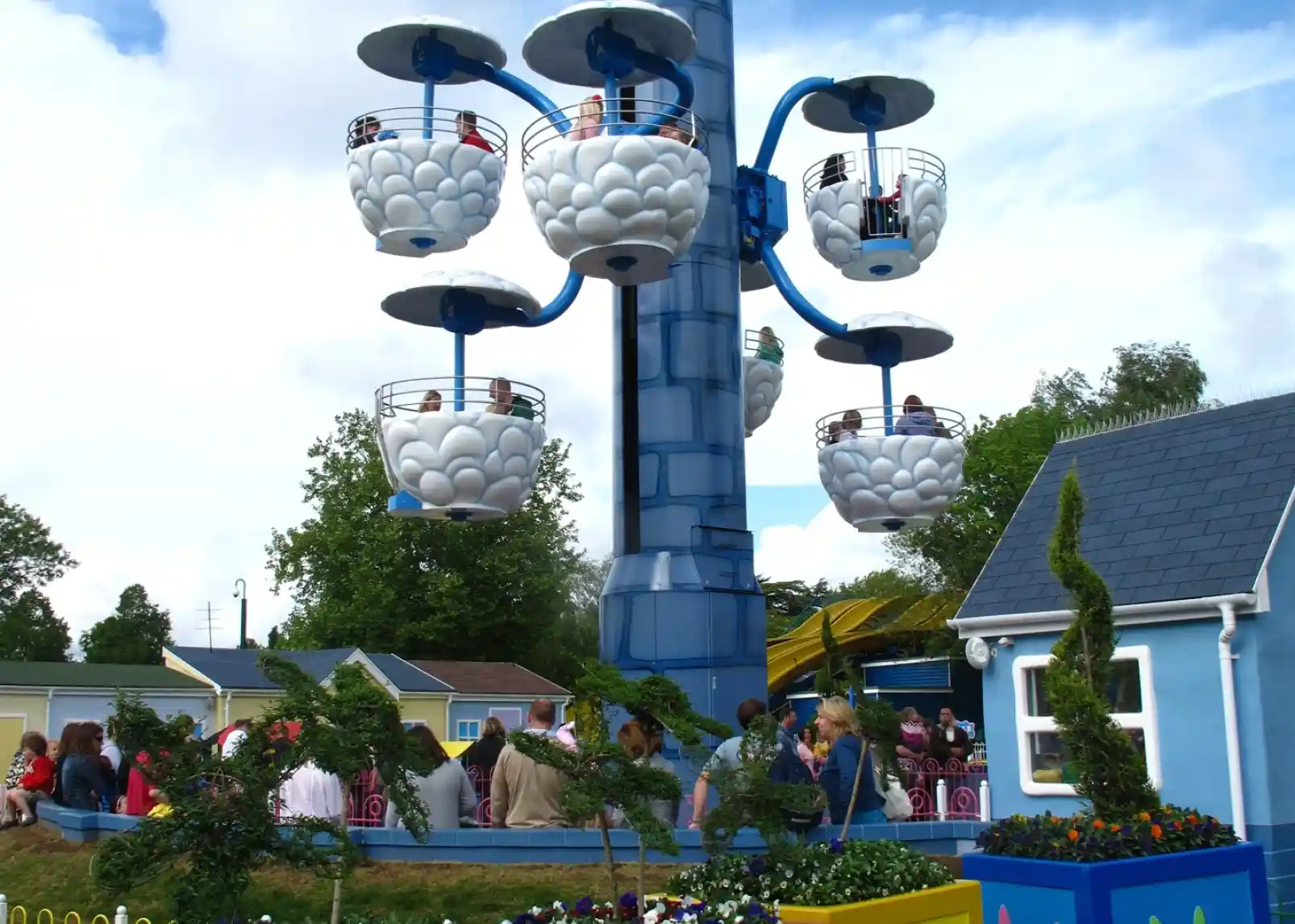 A colorful amusement park ride resembling oversized teacups hangs from a tall blue structure. People are seated inside the cups, enjoying the ride. In the foreground, vibrant flower arrangements and creative topiaries decorate the area, with families gathered nearby. The sky is partly cloudy, adding to the cheerful atmosphere.