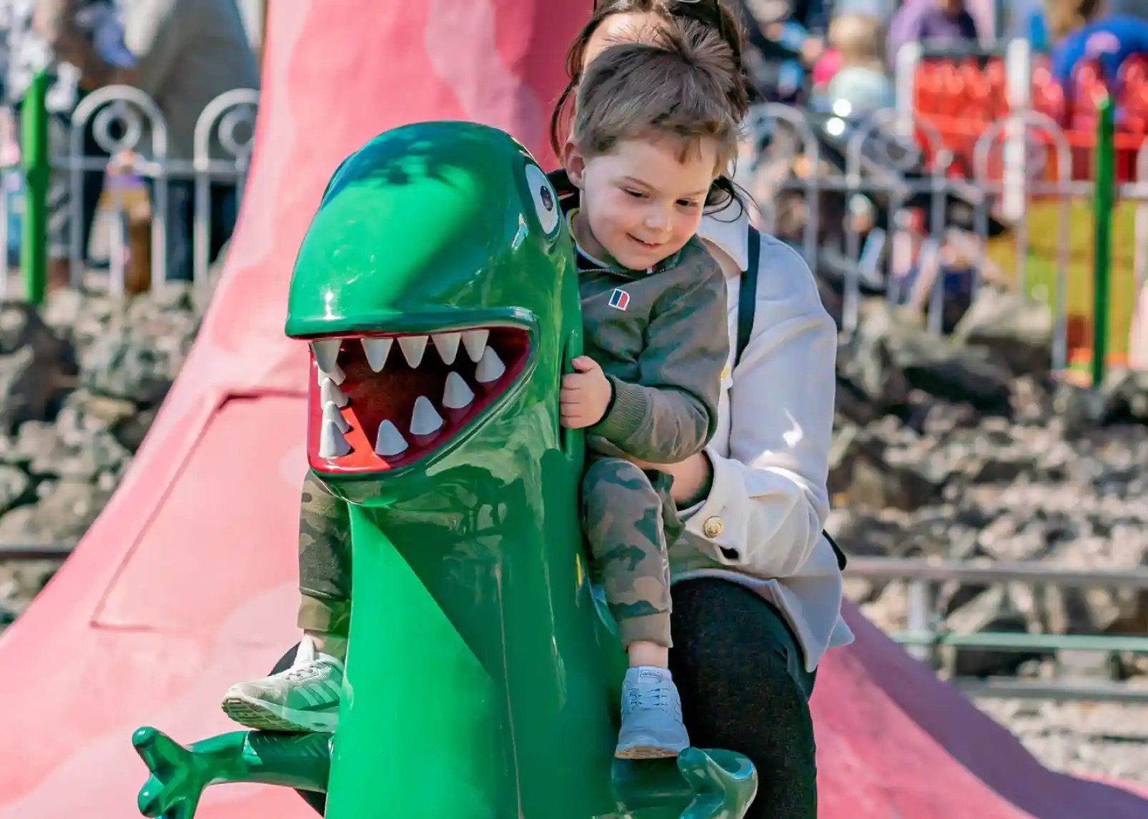 A young boy with short brown hair smiles as he rides a green dinosaur-shaped playground toy, while an adult woman behind him gently holds onto him. The background shows a colorful playground filled with other children and structures.