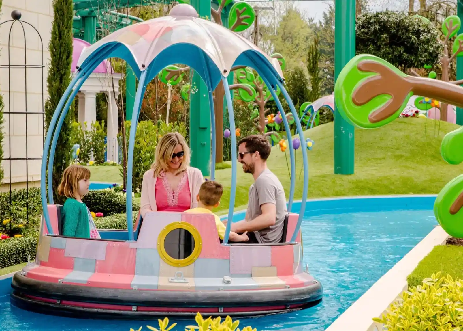 A family enjoys a colorful ride in a circular boat on a water attraction, surrounded by lush greenery and whimsical scenery. The mother and father sit with two children, smiling and interacting as they navigate the cheerful environment.