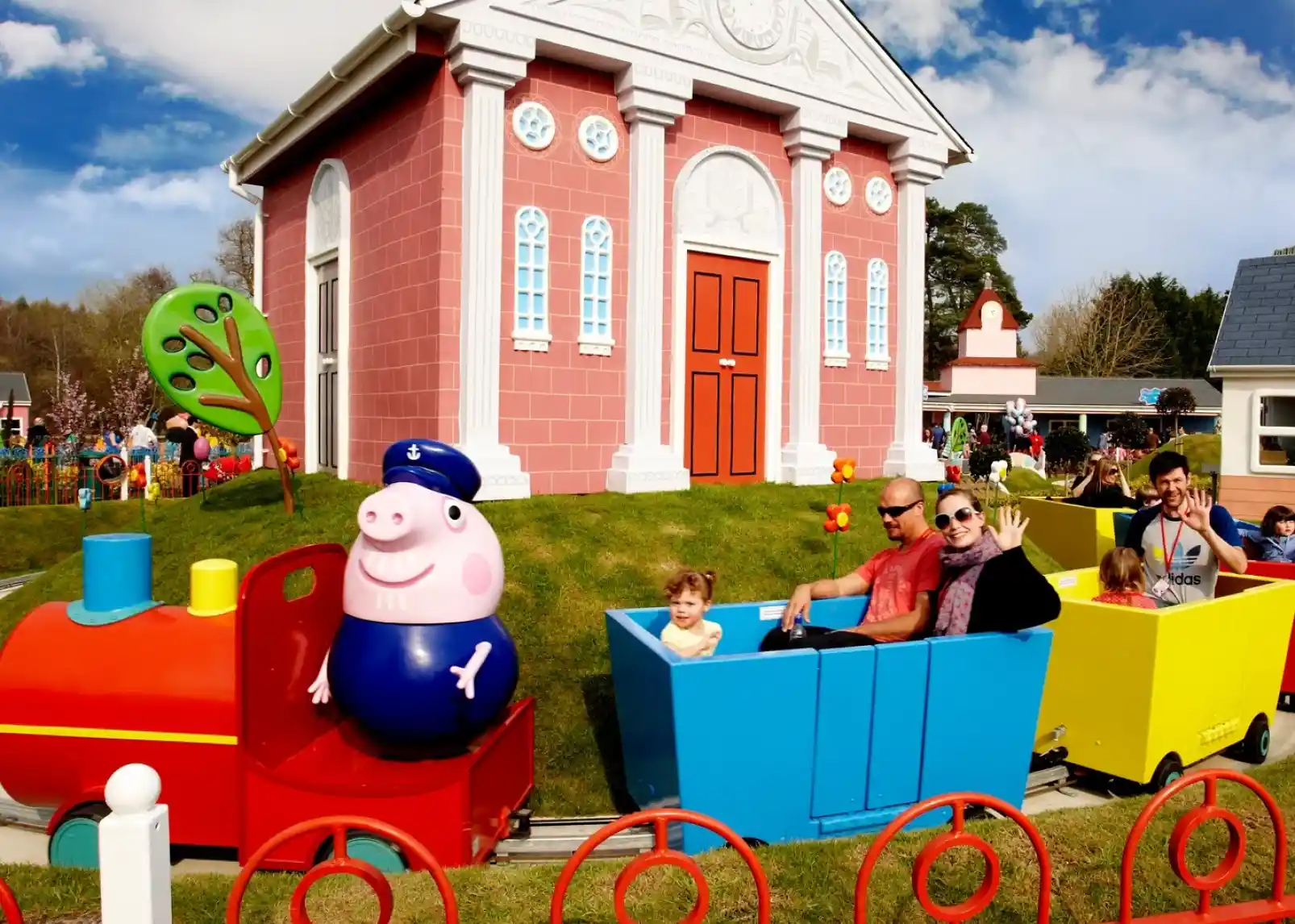 A colorful train ride styled like a cartoon, featuring a smiling pig character. Families are enjoying the ride, with one child sitting next to an adult in a red train car and others in bright blue and yellow cars. A pink building serves as a backdrop under a partly cloudy sky.