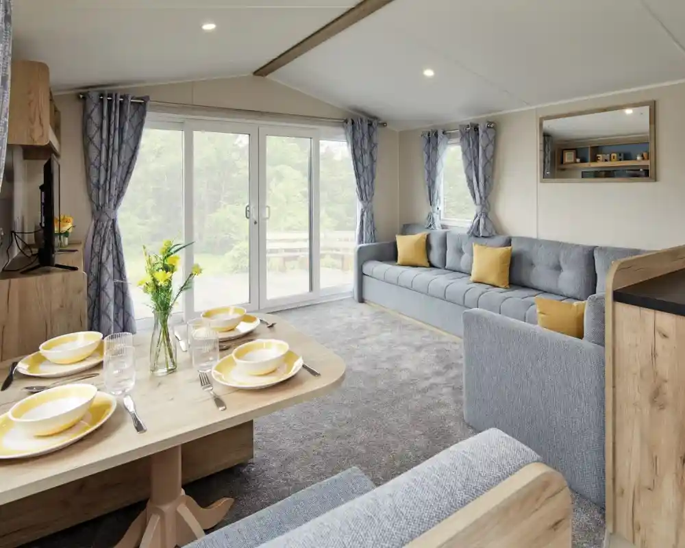 A modern living space featuring a gray sofa adorned with yellow cushions, a light wooden dining table set for four with yellow plates, and large glass doors that open to a patio area. Curtains with a blue pattern frame the windows, and there are fresh flowers on the table.