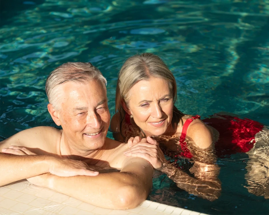 An elderly couple enjoys time together in a swimming pool, smiling and relaxed, with sunlight reflecting off the water.