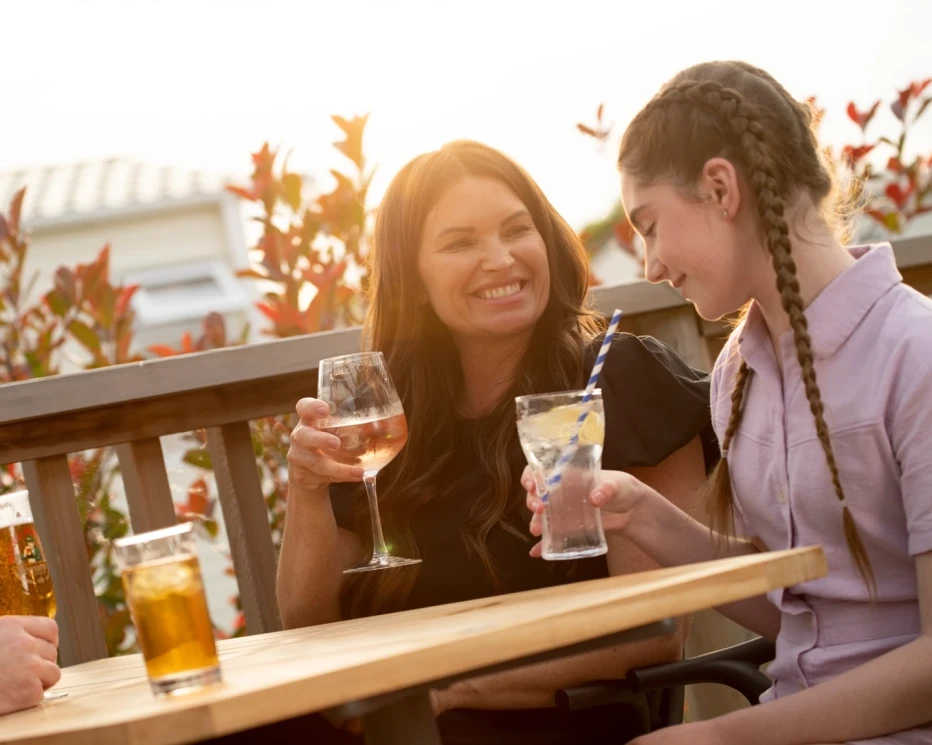 A woman and a girl smile at each other while seated at an outdoor table. The woman holds a glass of rosé wine, and the girl has a drink with a straw. Soft sunlight illuminates the scene, with greenery in the background.