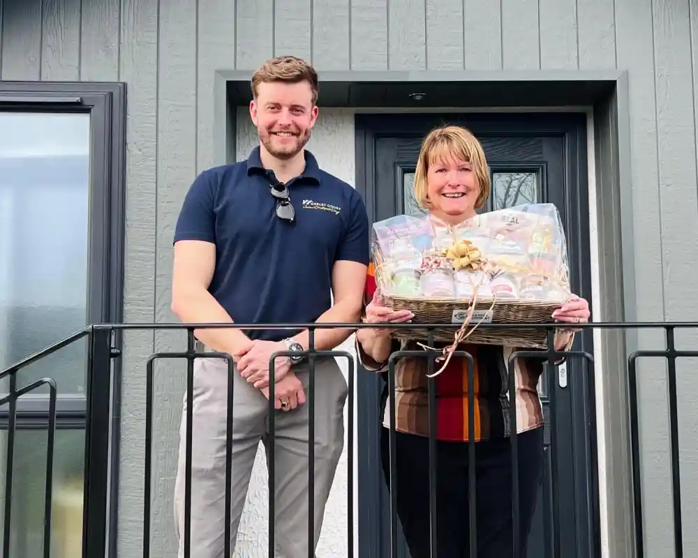 A man and a woman stand on a balcony outside a house, smiling at the camera. The woman holds a large gift basket filled with various goodies, while the man stands beside her with an arm resting on the railing. The house features a modern gray exterior.