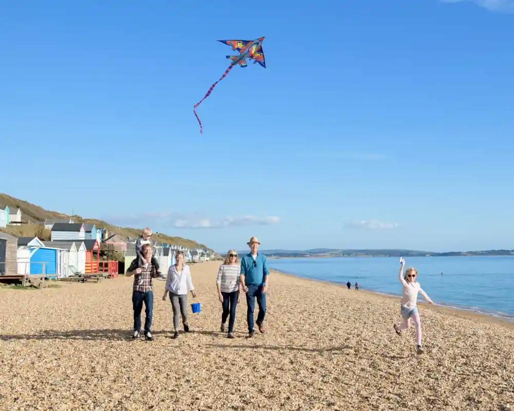 A family walks along a pebble beach, enjoying a sunny day. One child runs joyfully while another flies a colorful kite in the sky. Beach huts are visible in the background, and the calm sea extends towards the horizon.
